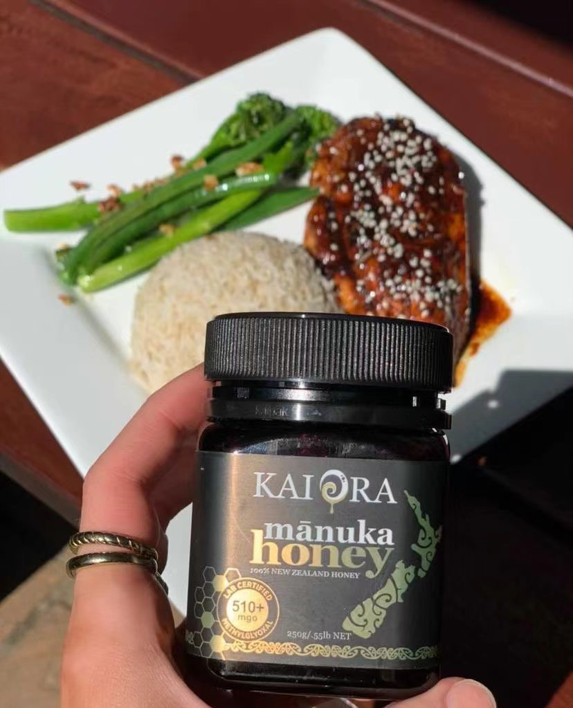 Kai Ora 510+ MGO mānuka honey jar held in front of a plate of sesame-crusted honey soy salmon with jasmine rice and garlic broccolini
